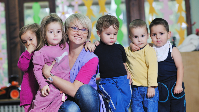 An image of a middle-aged, caucasian woman with a group of 5 preschool students
